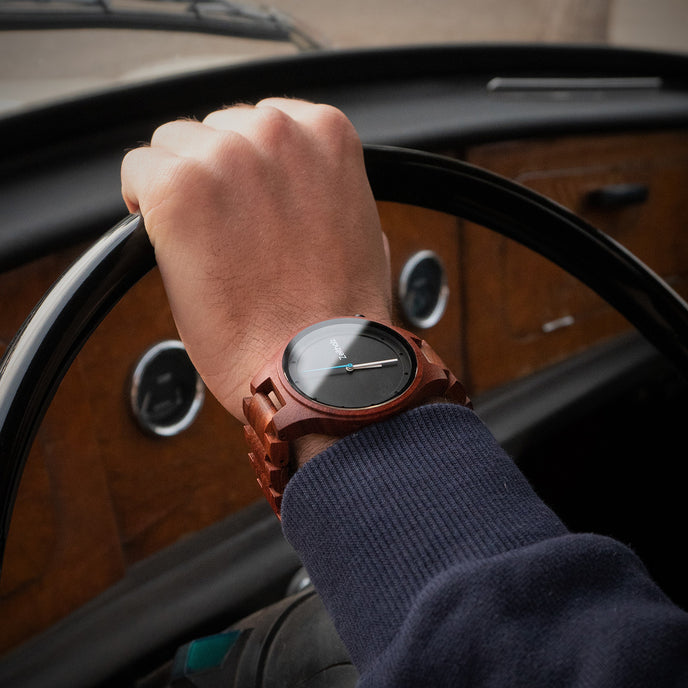 Männliches Handgelenk mit einer Zeitholz Holzuhr vor dem Lenkrad eines klassischen Autos mit Holzarmaturenbrett.